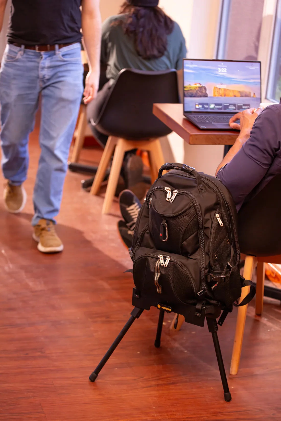 A backpack held upright on the Ryzer Go beside café tables where people work on laptops